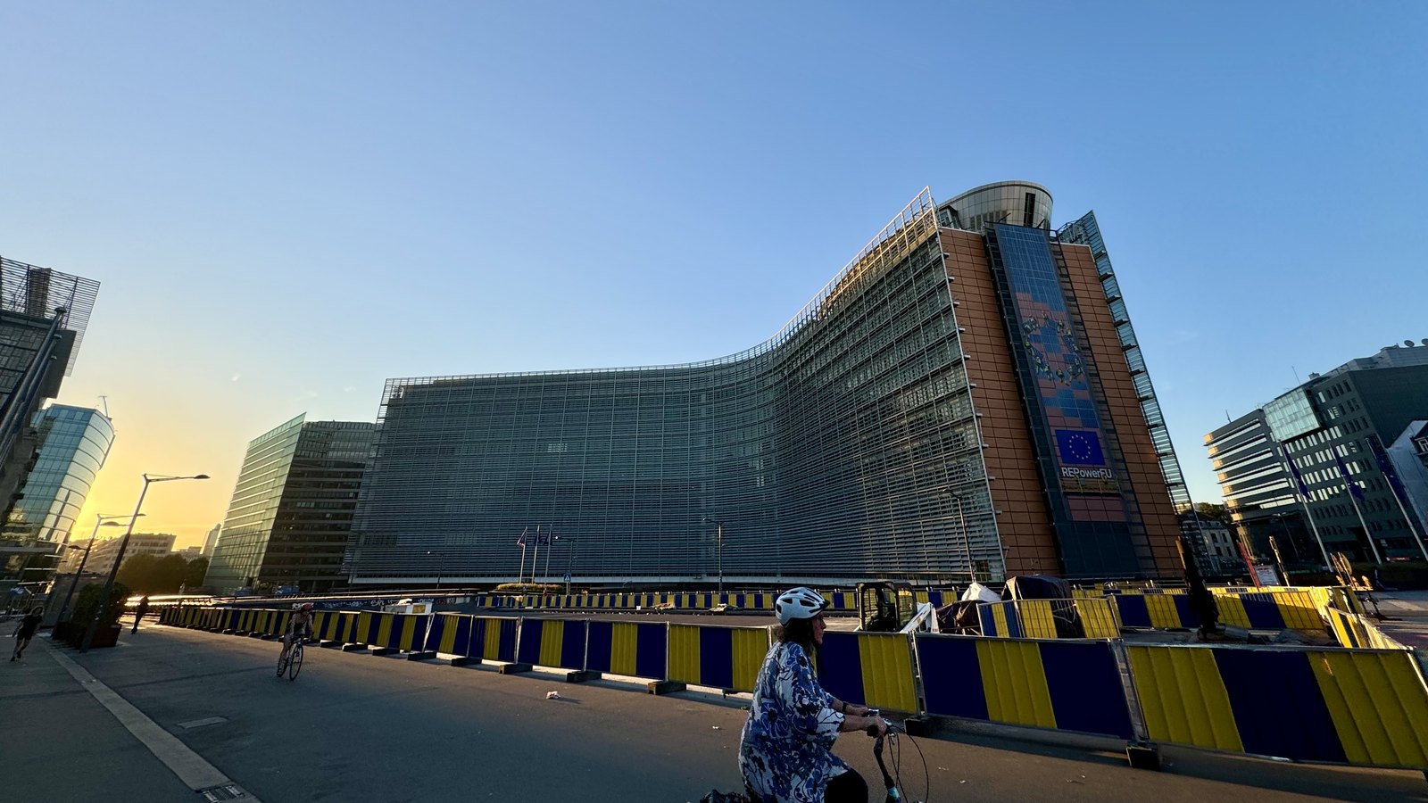 Le bâtiment du Berlaymont, siège de la Commission européenne à Bruxelles, en fin de journée sous un ciel bleu, avec des barrières bleues et jaunes au premier plan et une cycliste