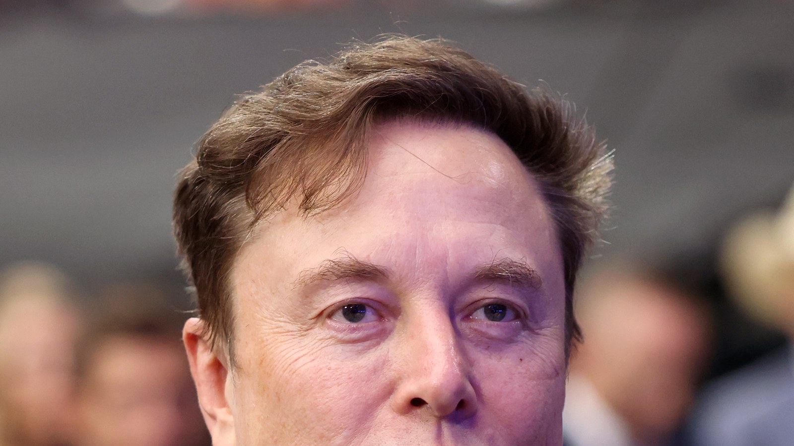 Portrait d'Elon Musk en gros plan, le regard tourné vers la droite, devant un public flou
