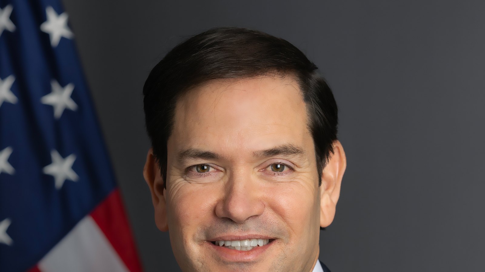 Portrait officiel de Marco Rubio, secrétaire d'État des États-Unis, souriant en costume sombre devant un drapeau américain