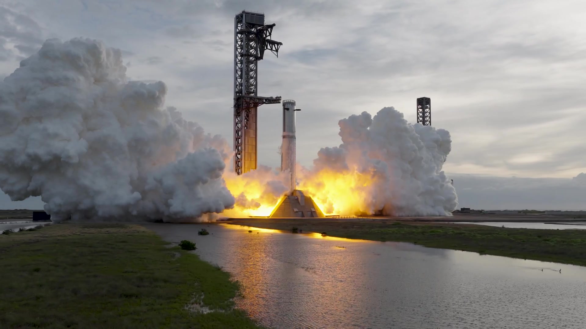 Le booster Super Heavy de SpaceX allume ses 33 moteurs sur le pas de tir de Starbase, entouré de nuages de vapeur, à côté de la tour de lancement