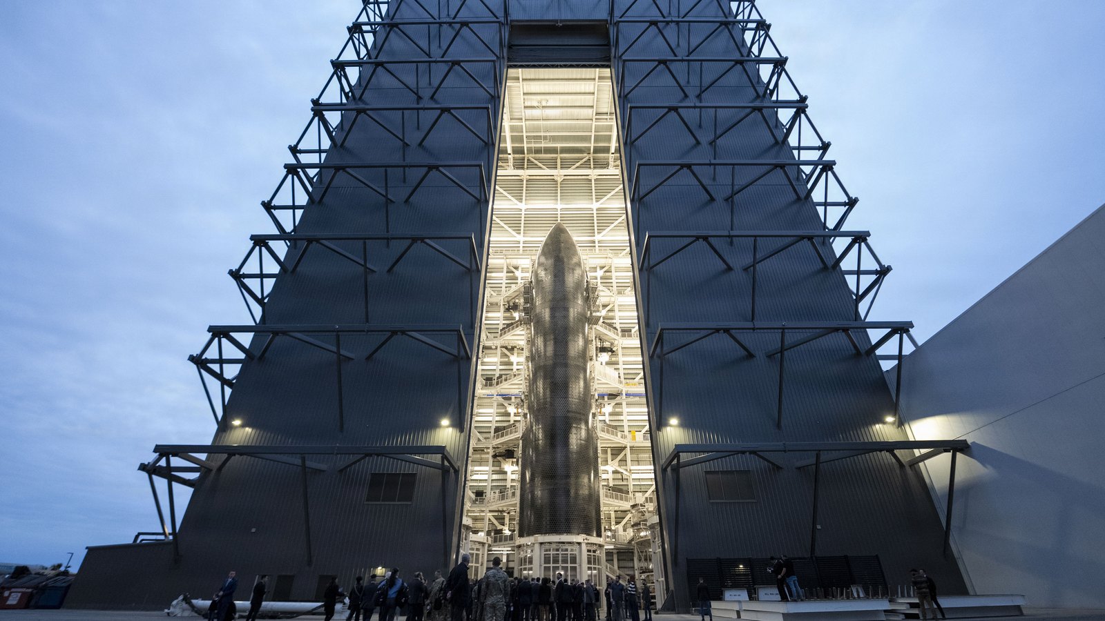 Grand hall d'assemblage de SpaceX à Starbase, au Texas, porte ouverte et éclairée sur un vaisseau Starship, au crépuscule, avec un groupe de visiteurs au pied du bâtiment