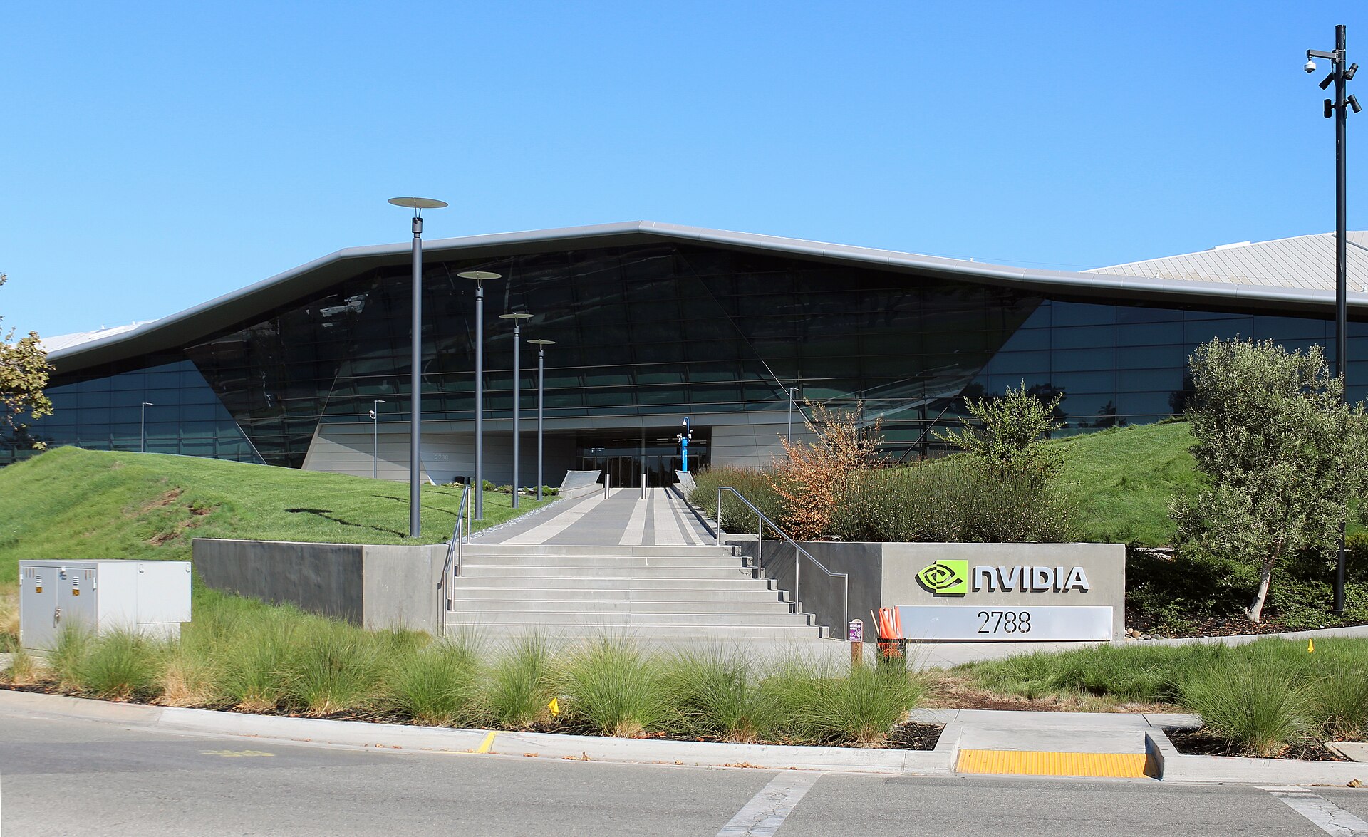 Entrée du siège de Nvidia à Santa Clara : grand bâtiment vitré au toit triangulaire, escalier entre deux talus gazonnés et panneau au logo Nvidia, sous un ciel bleu