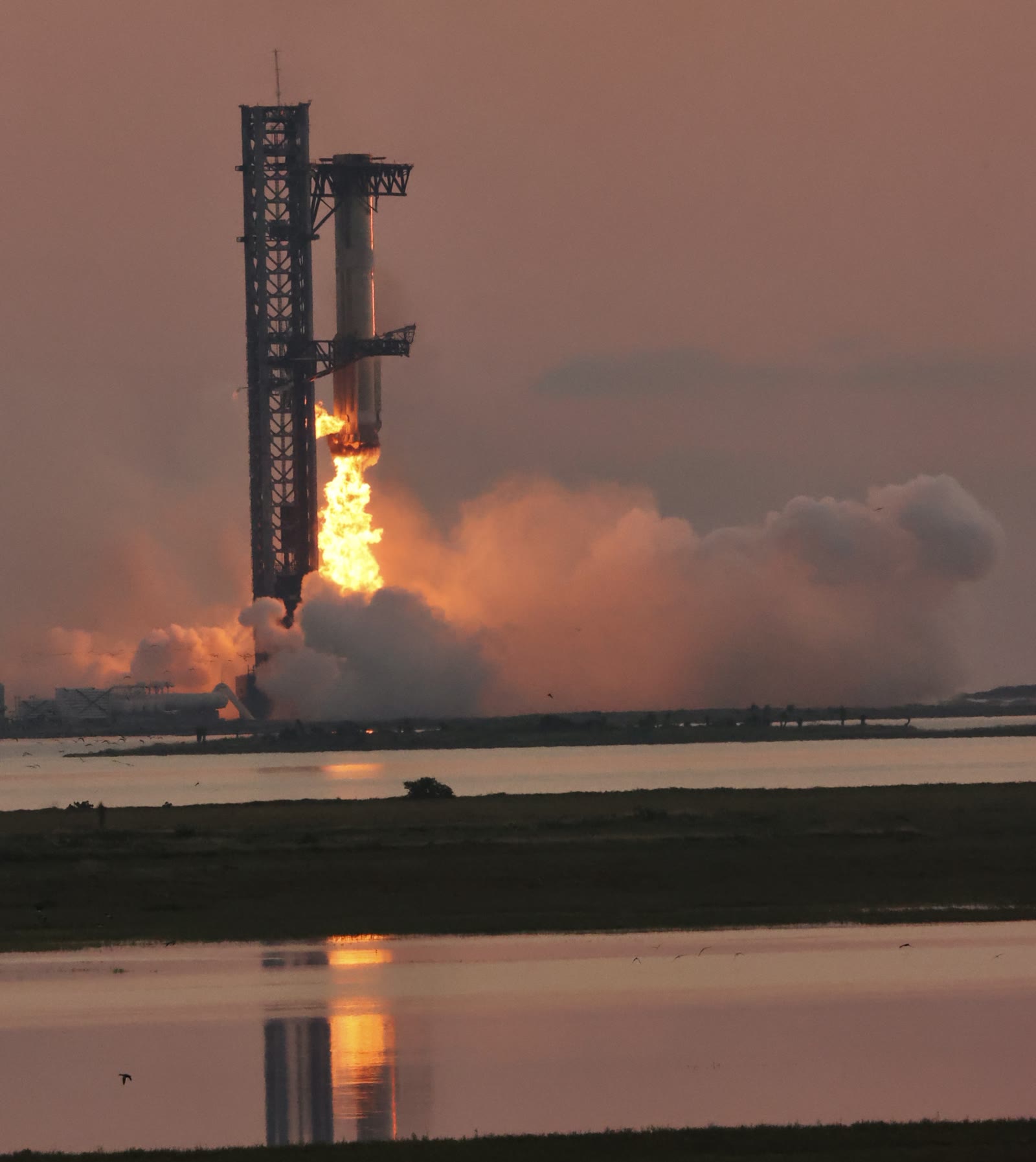 Le booster Super Heavy de SpaceX tenu par les bras de la tour de lancement de Starbase, moteurs encore allumés, au coucher du soleil
