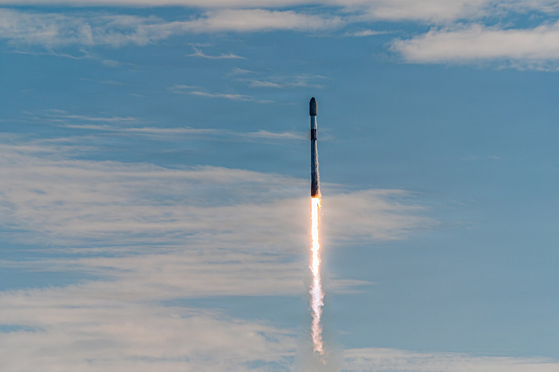 Une fusée Falcon 9 s'élève dans un ciel bleu voilé de nuages, laissant derrière elle une longue flamme blanche