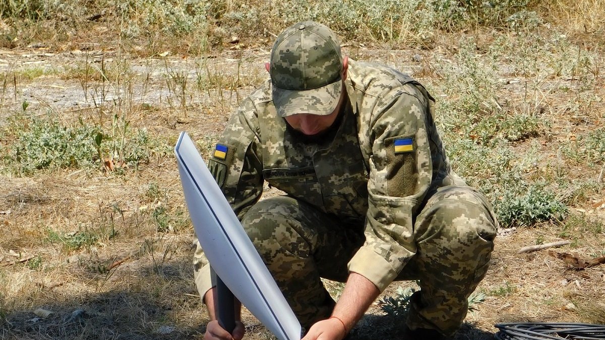 Un soldat ukrainien en tenue de camouflage, accroupi dans l'herbe sèche, fixe une antenne Starlink blanche sur son support