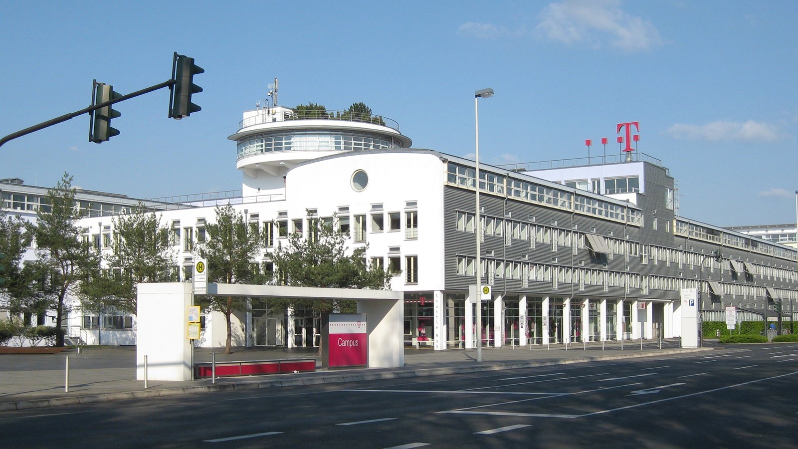 Bâtiments blancs et gris du campus de Deutsche Telekom à Bonn, avec le logo T magenta sur le toit, un abri de bus et des feux de circulation au premier plan, sous un ciel bleu