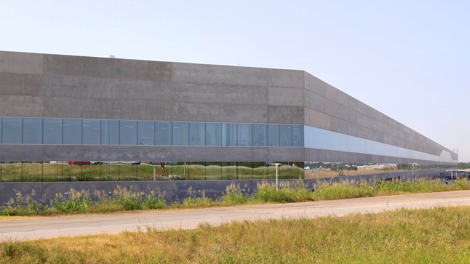 Façade grise et vitrée du bâtiment principal de la Gigafactory Texas de Tesla à Austin, au bord d'une route bordée d'herbes