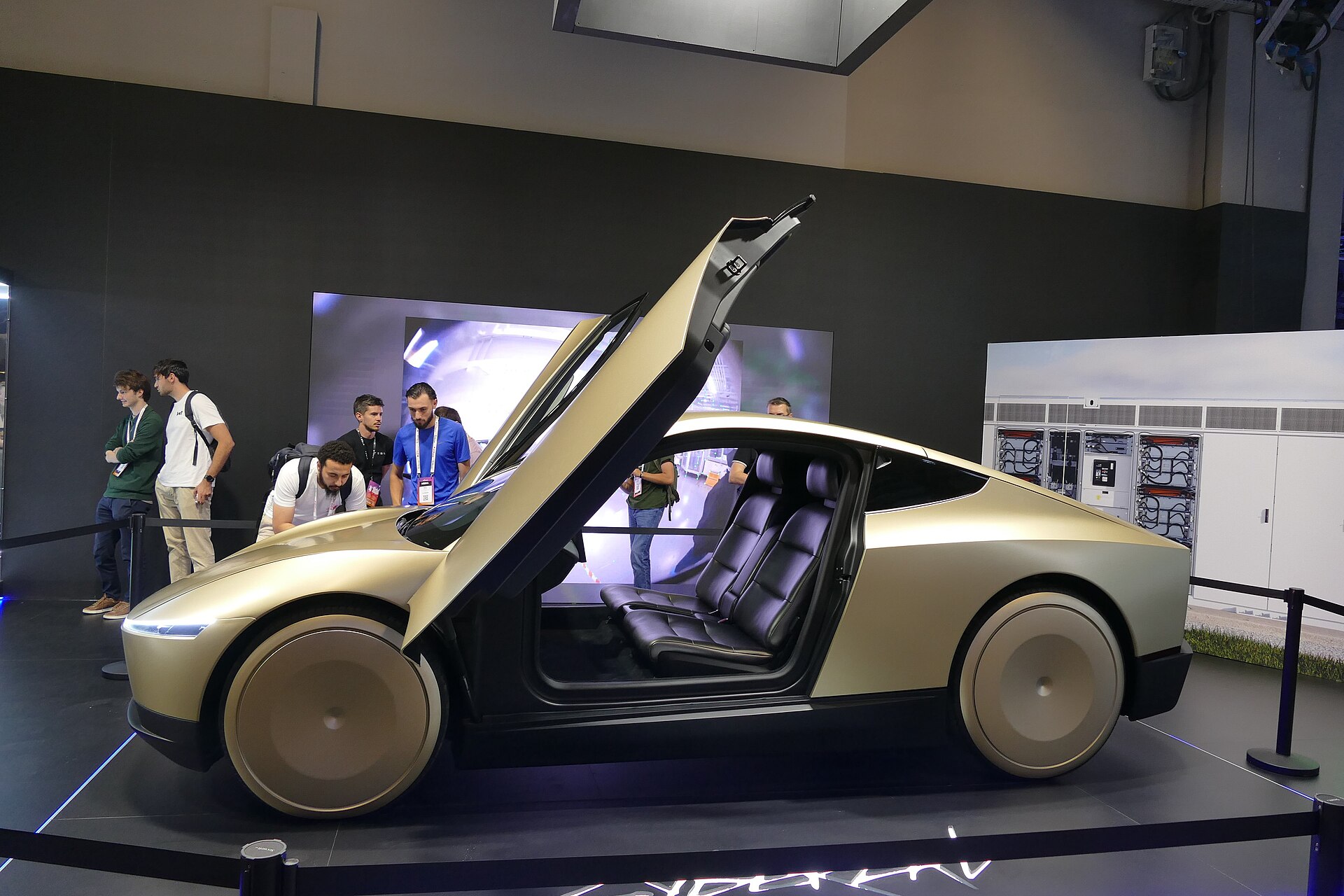 Un Cybercab doré de Tesla, porte papillon ouverte sur deux sièges, exposé sur un stand devant des visiteurs