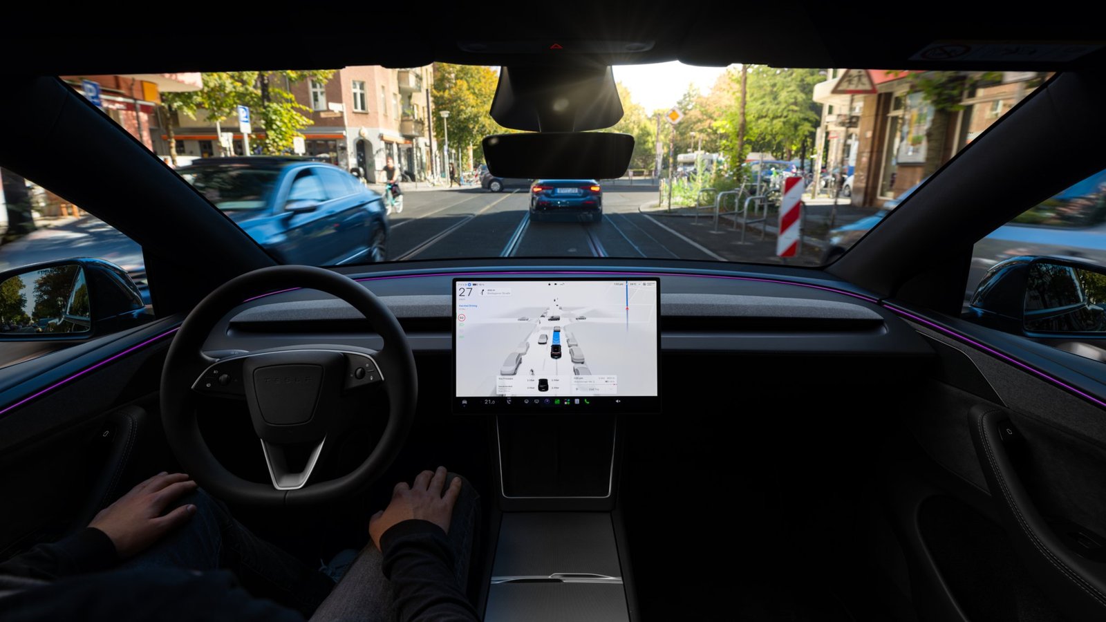 Vue depuis l'habitacle d'une Tesla qui roule dans une rue avec des rails de tram, mains du conducteur posées sur les genoux, écran central affichant la visualisation de l'aide à la conduite