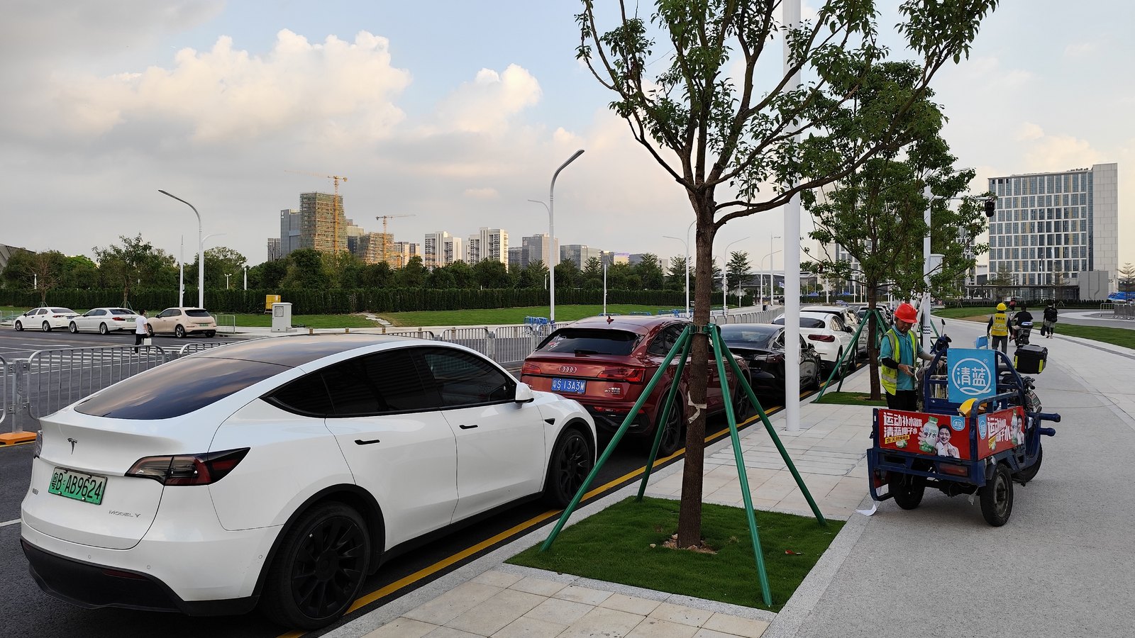 Tesla Model Y blanche garée le long d'un trottoir planté d'arbres à Shenzhen, devant d'autres voitures, avec des immeubles en construction à l'horizon