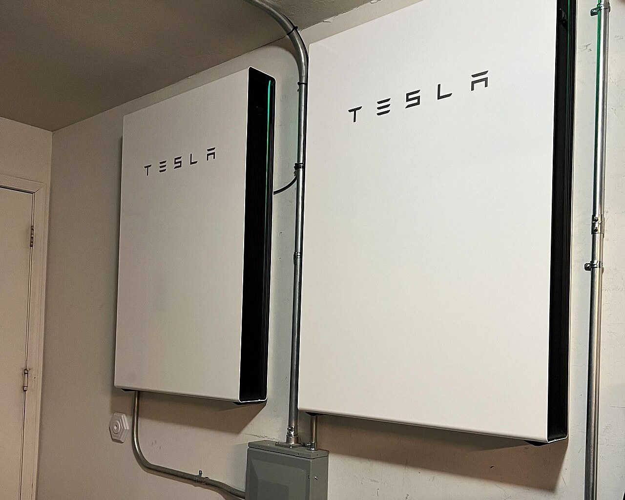 Deux batteries murales blanches Tesla Powerwall 3 fixées au mur d'un garage, reliées par des gaines électriques à un boîtier de coupure gris